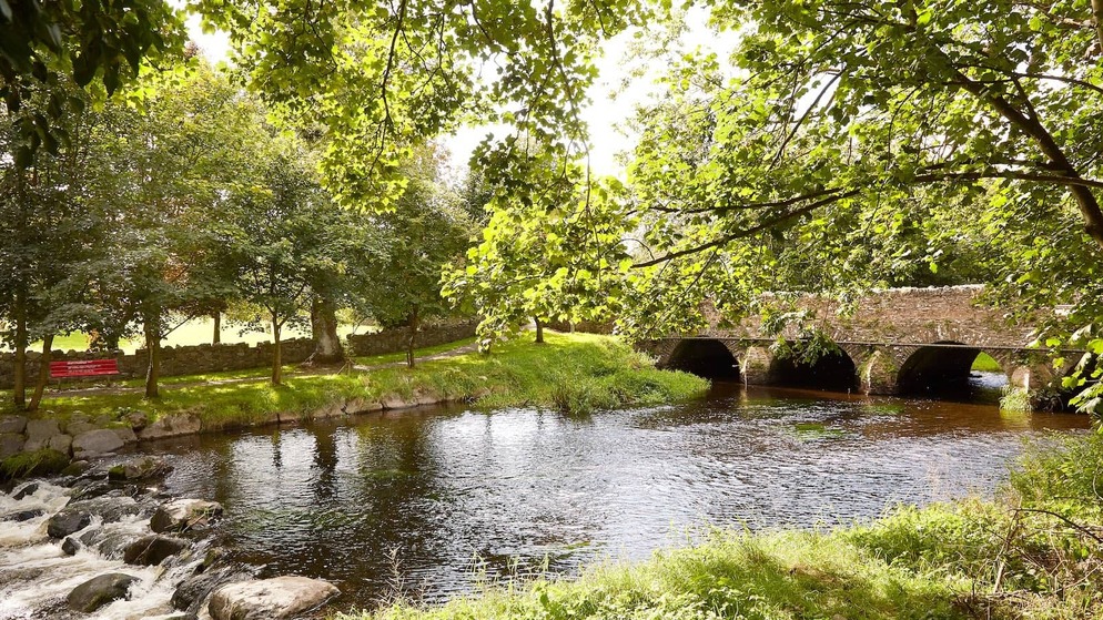 Sunlit view of the River Fane in Inniskeen, County Monaghan, with leafy trees and a stone arch bridge over calm water.