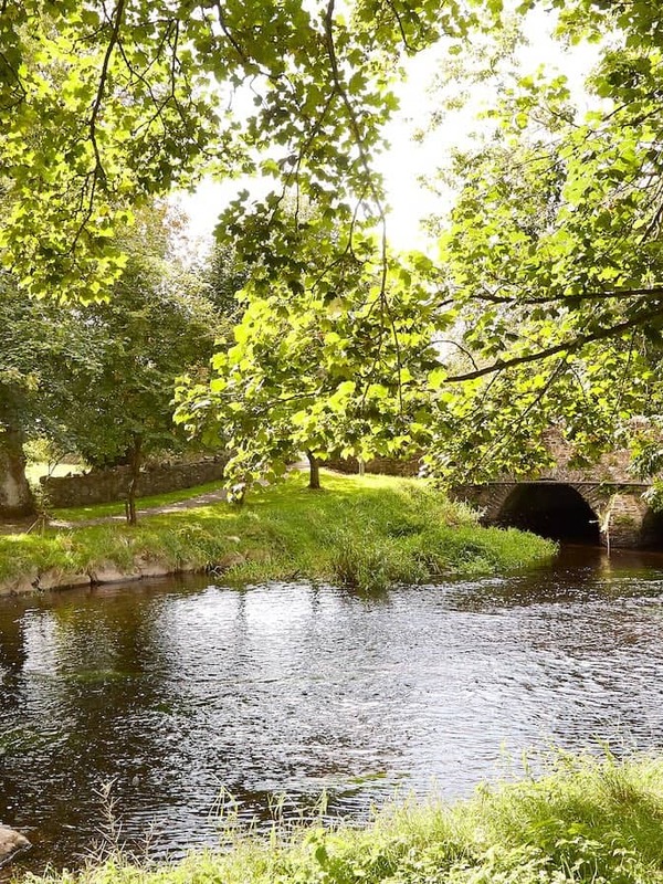 Sunlit view of the River Fane in Inniskeen, County Monaghan, with leafy trees and a stone arch bridge over calm water.