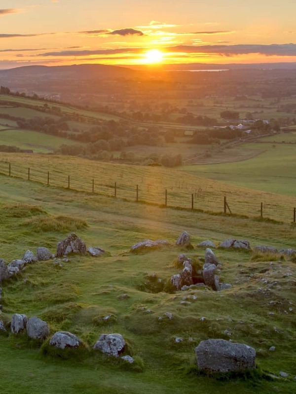 Sunset over the Loughcrew Cairns in County Meath, with a stone circle on grassy hills overlooking patchwork farmland.