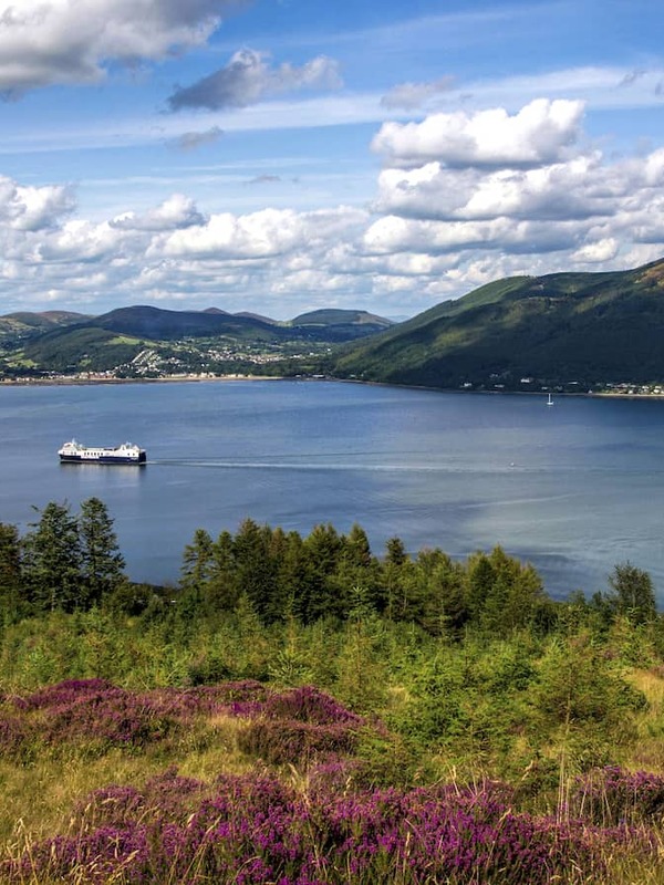 View over Carlingford Lough with a ferry crossing calm water, framed by heather-covered hills and distant mountains.