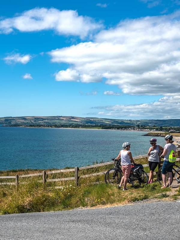 Cyclists on the Waterford Greenway stopping to admire coastal views across a calm bay under a bright blue sky.