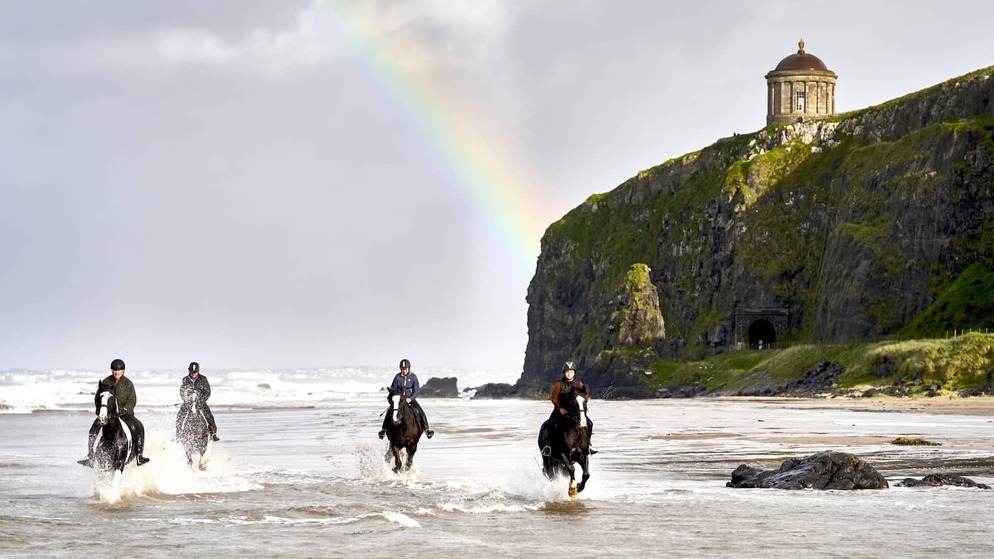 A group horse riding on a beach in County Londonderry with a rainbow and a clifftop temple in the background.