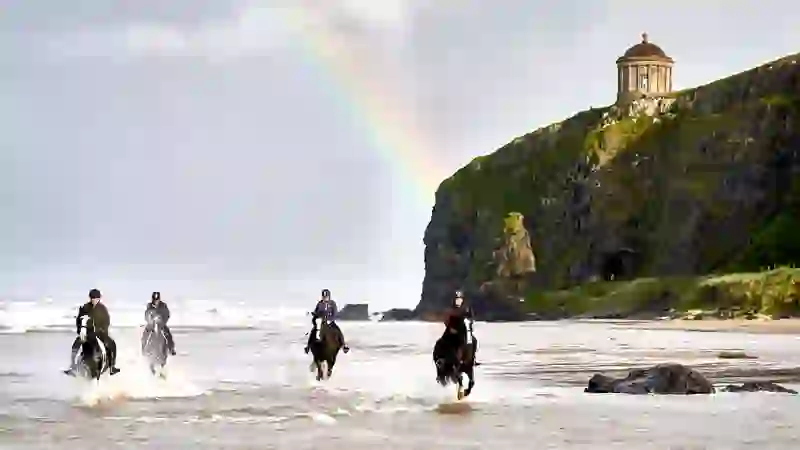 A group horse riding on a beach in County Londonderry with a rainbow and a clifftop temple in the background.