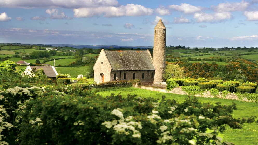 Stone church with a round tower surrounded by hedgerows and wildflowers in the green countryside of County Down.