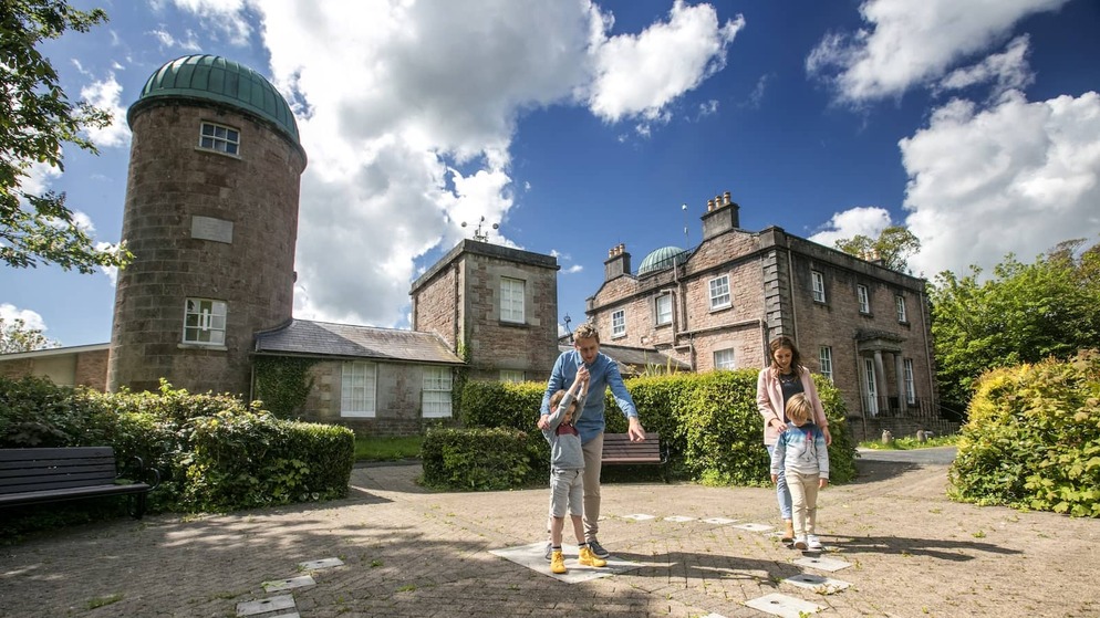 Family exploring the grounds of Armagh Observatory with domed tower and heritage buildings under a bright sky.
