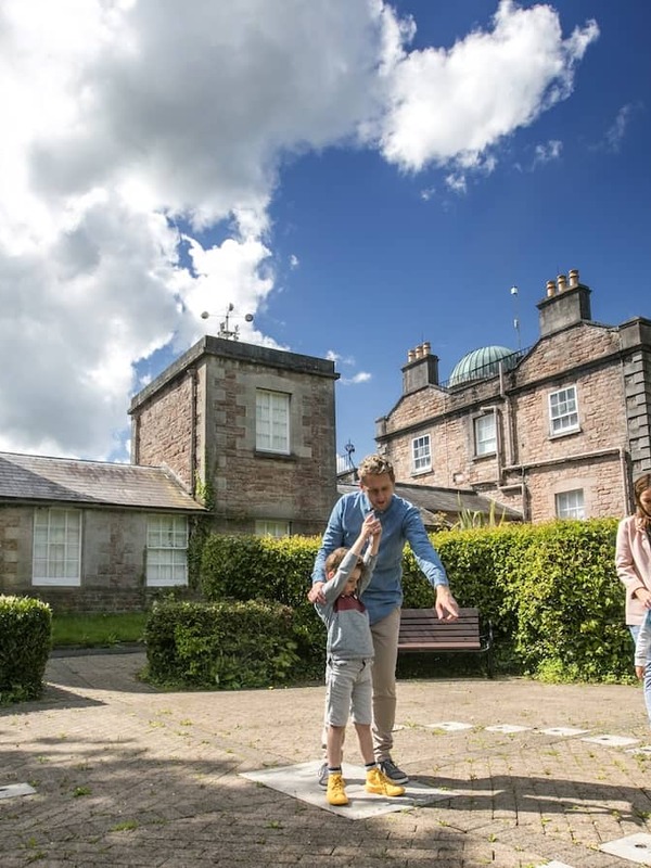 Family exploring the grounds of Armagh Observatory with domed tower and heritage buildings under a bright sky.