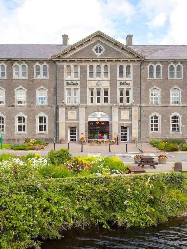 Grand stone building of Belleek Pottery framed by vibrant flowers and riverside greenery in County Fermanagh.