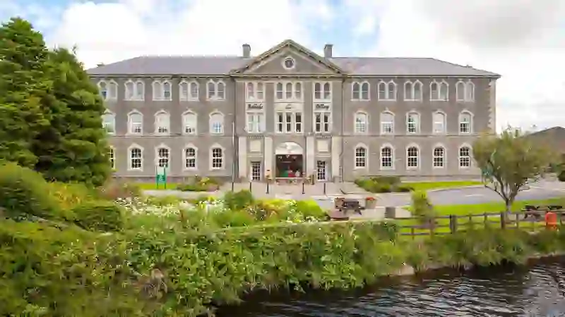 Grand stone building of Belleek Pottery framed by vibrant flowers and riverside greenery in County Fermanagh.