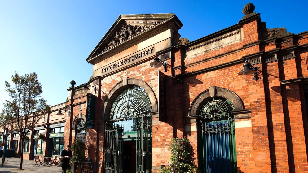 Red-brick Victorian facade of St George’s Market with arched gates and a café terrace in Belfast.