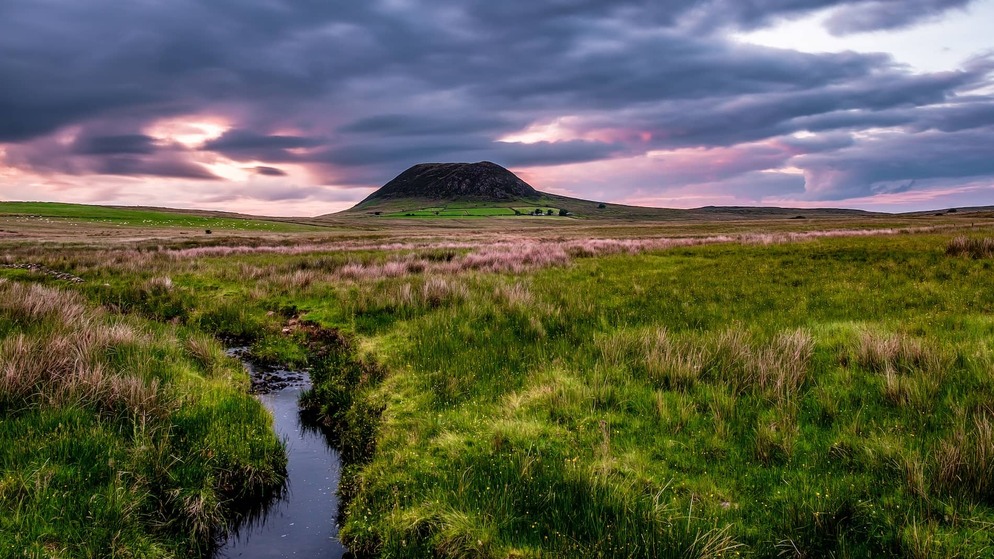 Slemish Mountain at sunset under dramatic clouds, with a winding stream and grassy moorland in County Antrim, Northern Ireland.