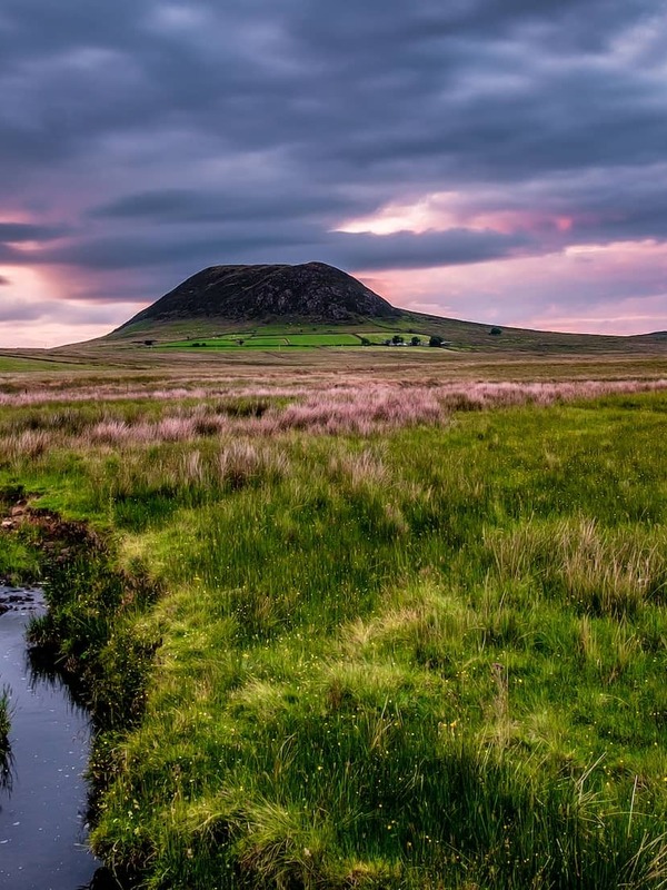 Slemish Mountain at sunset under dramatic clouds, with a winding stream and grassy moorland in County Antrim, Northern Ireland.