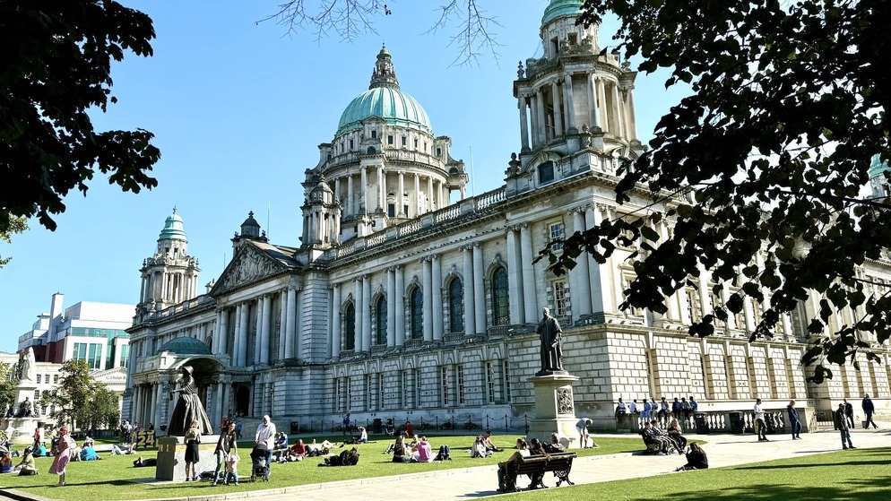 People relaxing on the lawn outside Belfast City Hall, a grand Baroque Revival building with green copper domes in Northern Ireland.