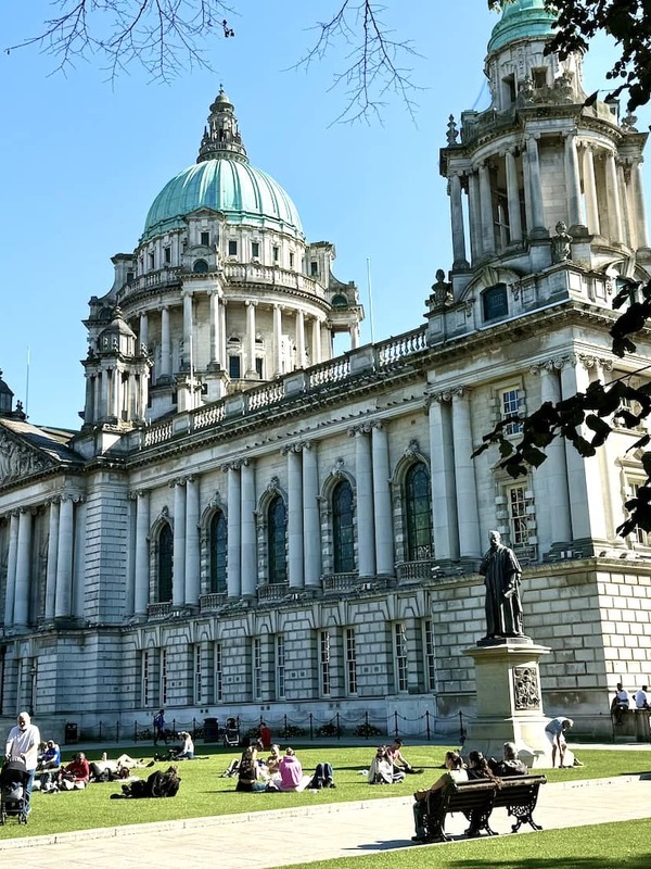 People relaxing on the lawn outside Belfast City Hall, a grand Baroque Revival building with green copper domes in Northern Ireland.