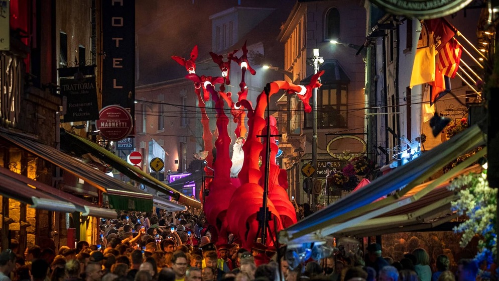Vibrant red creature-themed float moves through a bustling, lantern-lit street at the Galway International Arts Festival.