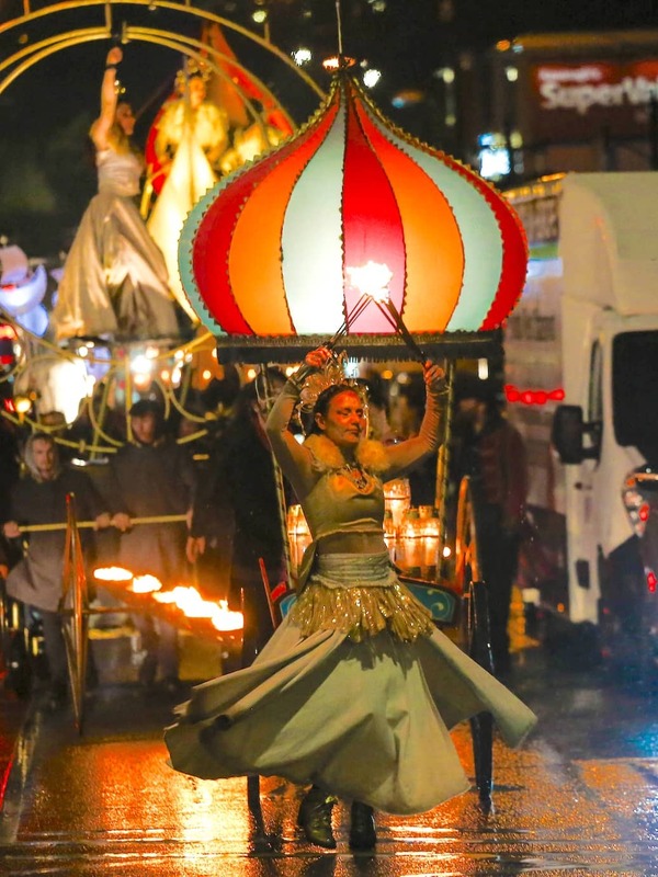 Performer twirling fire in a dramatic costume leads a night-time street parade with glowing floats and performers in flowing dresses.