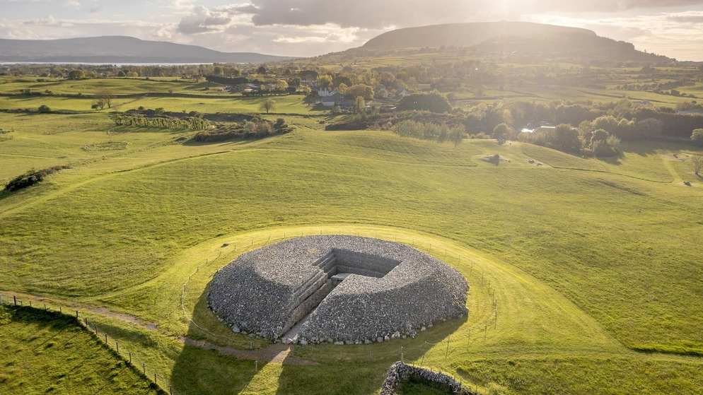 Aerial view of a large circular stone cairn at Carrowmore Megalithic Cemetery, set in green Sligo countryside with a glowing evening sky.