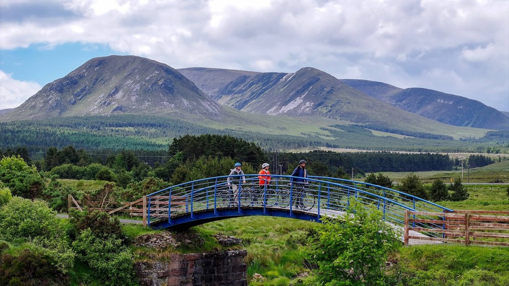 Cyclists cross a bridge on the Great Western Greenway in County Mayo with Nephin Beg mountains in the background.