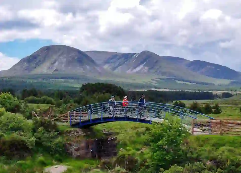 Cyclists cross a bridge on the Great Western Greenway in County Mayo with Nephin Beg mountains in the background.