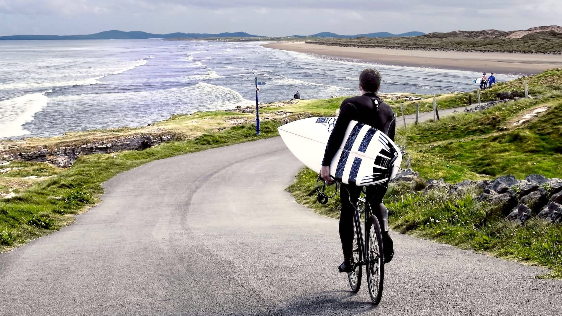 Surfer cycling with surfboard along scenic coastal road beside sandy beach in County Donegal.