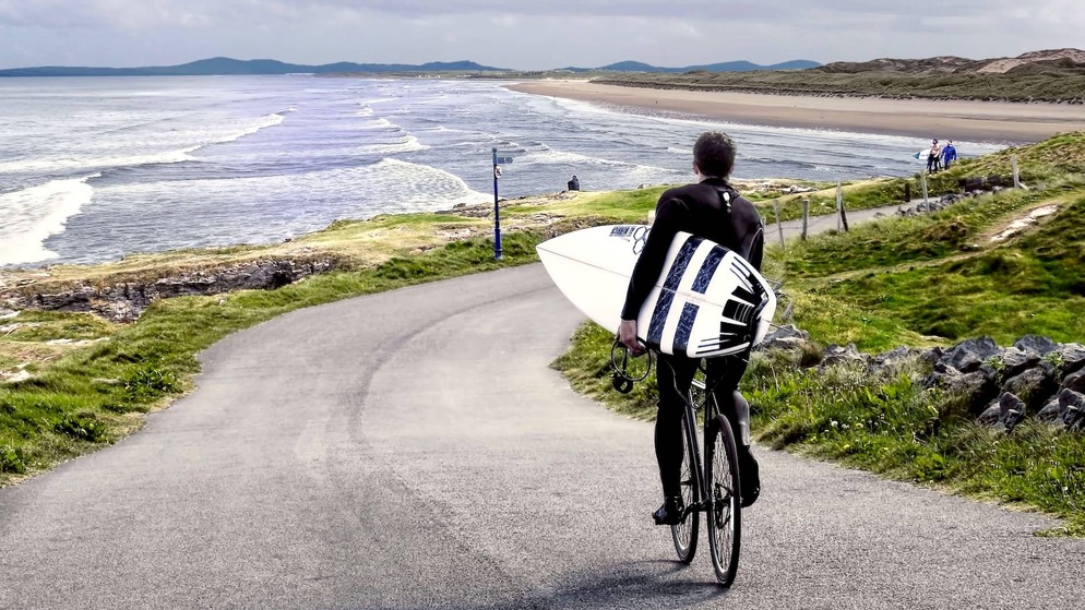 Surfer cycling with surfboard along scenic coastal road beside sandy beach in County Donegal.