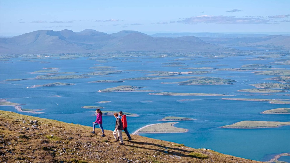 Three hikers walk above Clew Bay in County Mayo with views of its drumlin islands and distant mountains.