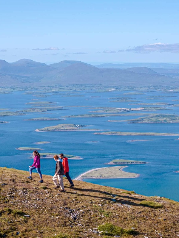 Three hikers walk above Clew Bay in County Mayo with views of its drumlin islands and distant mountains.