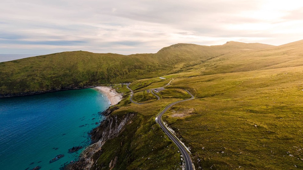 Winding cliff road leading to a golden beach and turquoise sea, backed by grassy hills at Keem Bay, Achill Island, County Mayo.