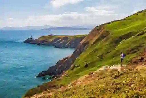 A hiker stands on a grassy cliff path overlooking the sea and lighthouse at Howth Head on a sunny day.