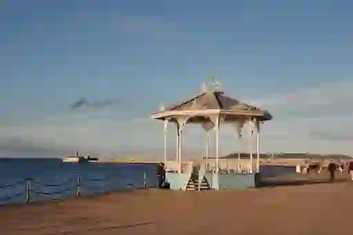 Victorian bandstand on Dún Laoghaire pier with distant lighthouse and walkers under clear evening light.