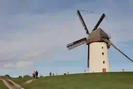 People walk a grassy hill past the historic Skerries Windmill under a soft blue sky with light cloud cover.