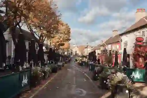 Outdoor dining terraces line a leafy street in Malahide village, with planters and historic shopfronts under autumn trees.