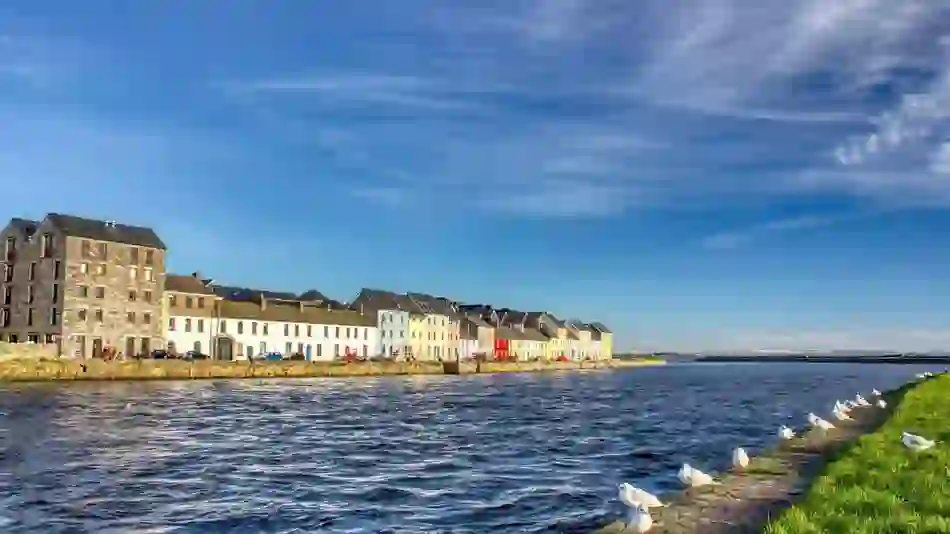Colourful houses along the Claddagh Quay waterfront in Galway City under a bright blue sky.