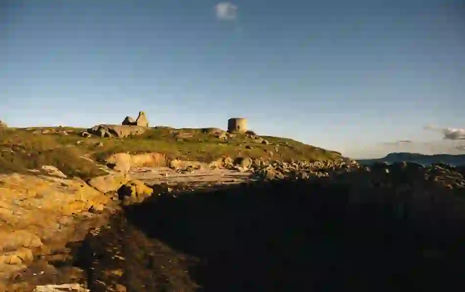 Martello tower and ruins on grassy Dalkey Island viewed from rocky shore with sea in foreground.