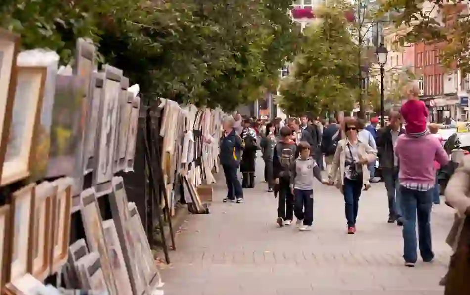 Crowds browsing outdoor art stalls with framed paintings along Dublin’s Merrion Square railings.