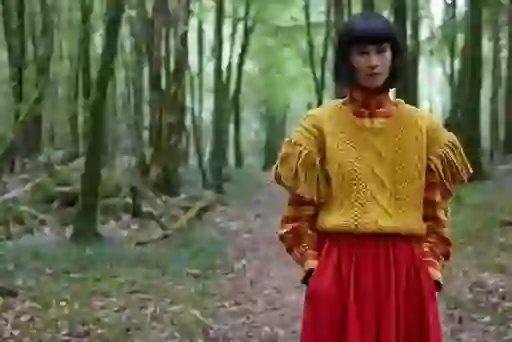 Model in forest path wearing a richly textured yellow wool sweater over a red plaid blouse and crimson skirt.