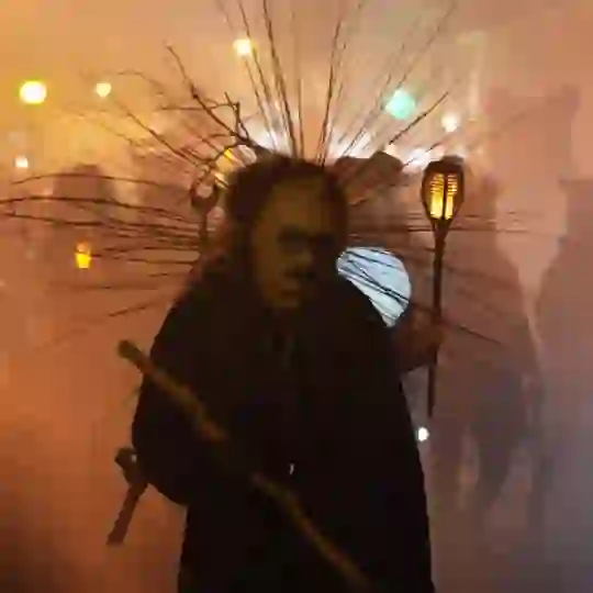 Costumed performer with spiked headdress and glowing lantern at an atmospheric festival procession.
