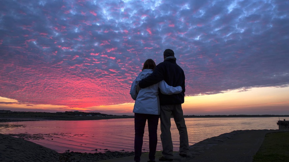 Couple standing arm in arm watching a vivid pink and purple sunset over calm waters off Burr Point, County Down.
