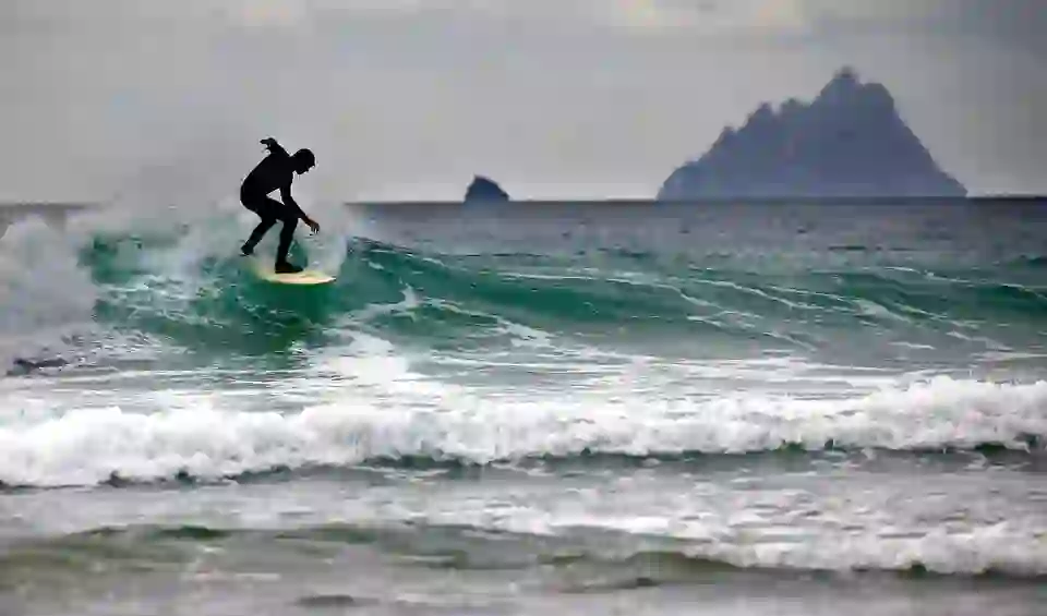 Surfer riding a wave off the Kerry coast with Skellig Michael island visible in the distance.
