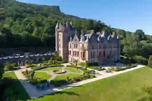 A grand Victorian castle with manicured gardens at the edge of a forested hillside.