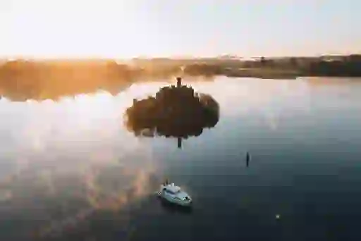 A castle ruin sits on a small wooded island surrounded by calm water and morning mist – a cruiser nearby..