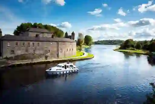 A cruiser boat passes a historic stone castle on a calm river beneath a blue sky.