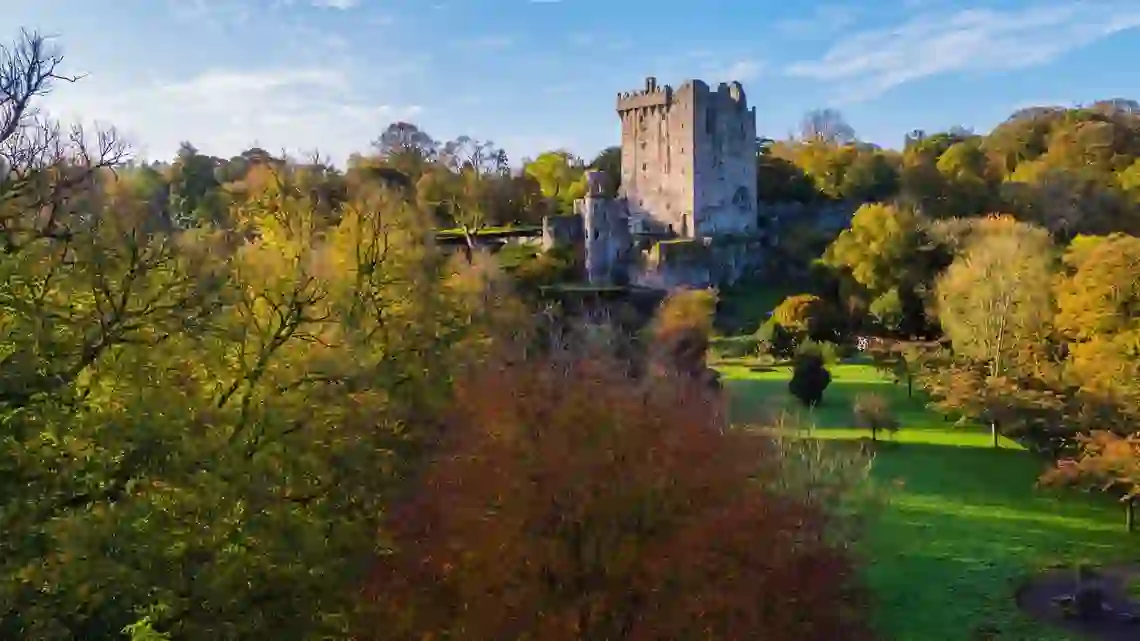 Blarney Castle surrounded by autumn trees and gardens in County Cork, Ireland, under a clear blue sky.