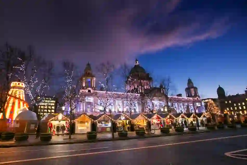 Belfast City Hall lit up at dusk with Christmas market stalls and festive lights in front.