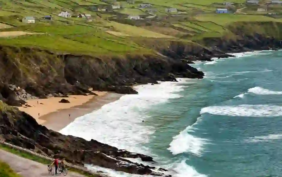 Coumeenole Beach on the Dingle Peninsula in County Kerry with waves hitting rocky cliffs and two cyclists on a coastal road.