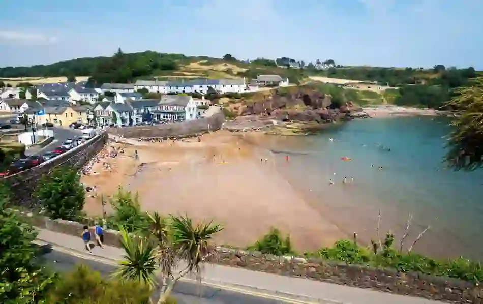 Dunmore East beach in County Waterford with people sunbathing and swimming near colourful buildings by the harbour.