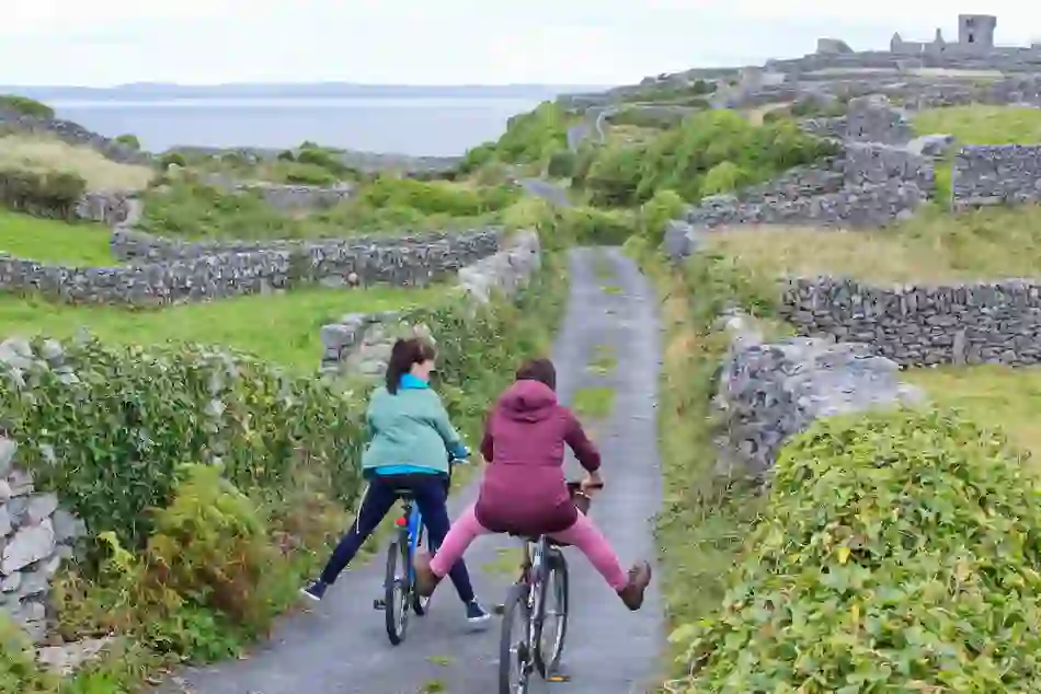 Two cyclists, one playfully with legs out, ride along a narrow path flanked by stone walls on the Aran Islands, Galway.