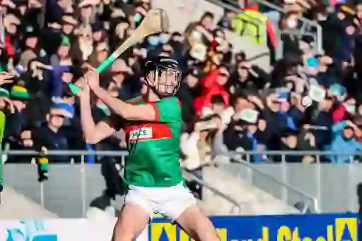 Hurling player in red and green kit swings hurley to strike sliotar during Cork senior final as crowd watches on.