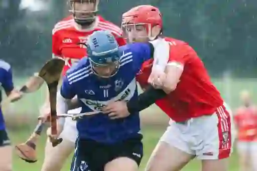 Player in blue jersey breaks past red-shirted defender during a wet hurling championship clash in Cork.