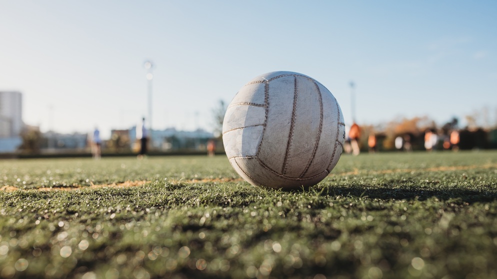 Gaelic football on sunlit pitch with blurred players training in the background at a GAA grounds.