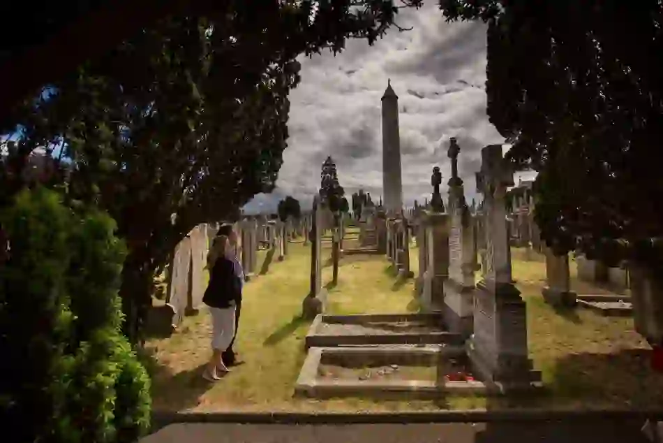 Visitors walking among historic gravestones at Glasnevin Cemetery, Dublin, with the tall round tower in the background.